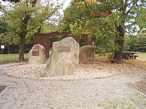 Drei große Gedenksteine umgeben von hohen Bäumen und Sitzmöglichkeiten. 